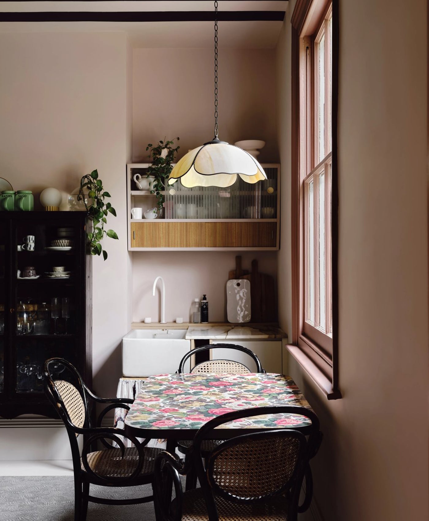 Plaster coloured dining area with floral tablecloth, wicker chairs, and a window with a lamp.