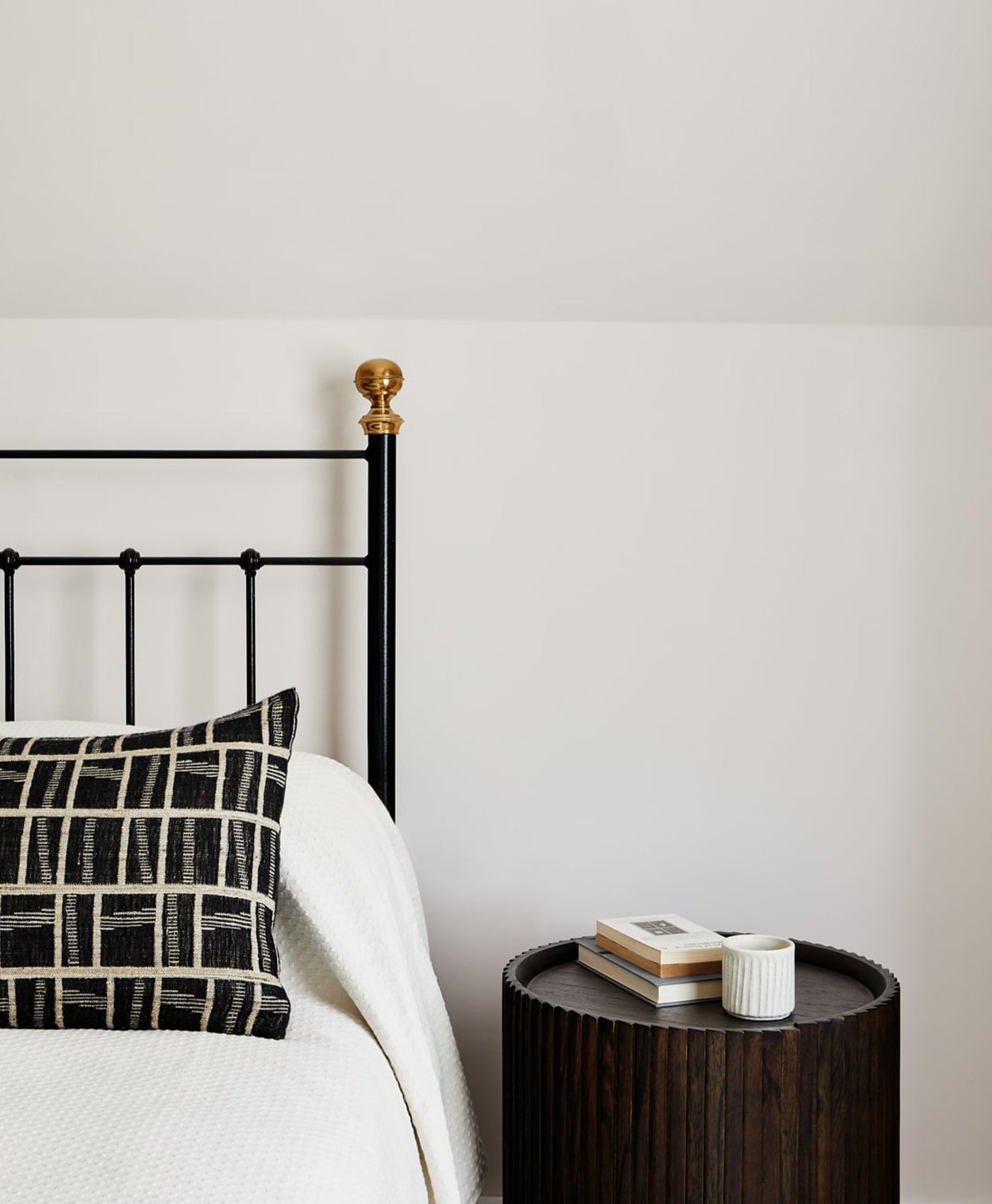 Bed with black metal headboard and off-white walls, next to a wooden side table with books and a mug.