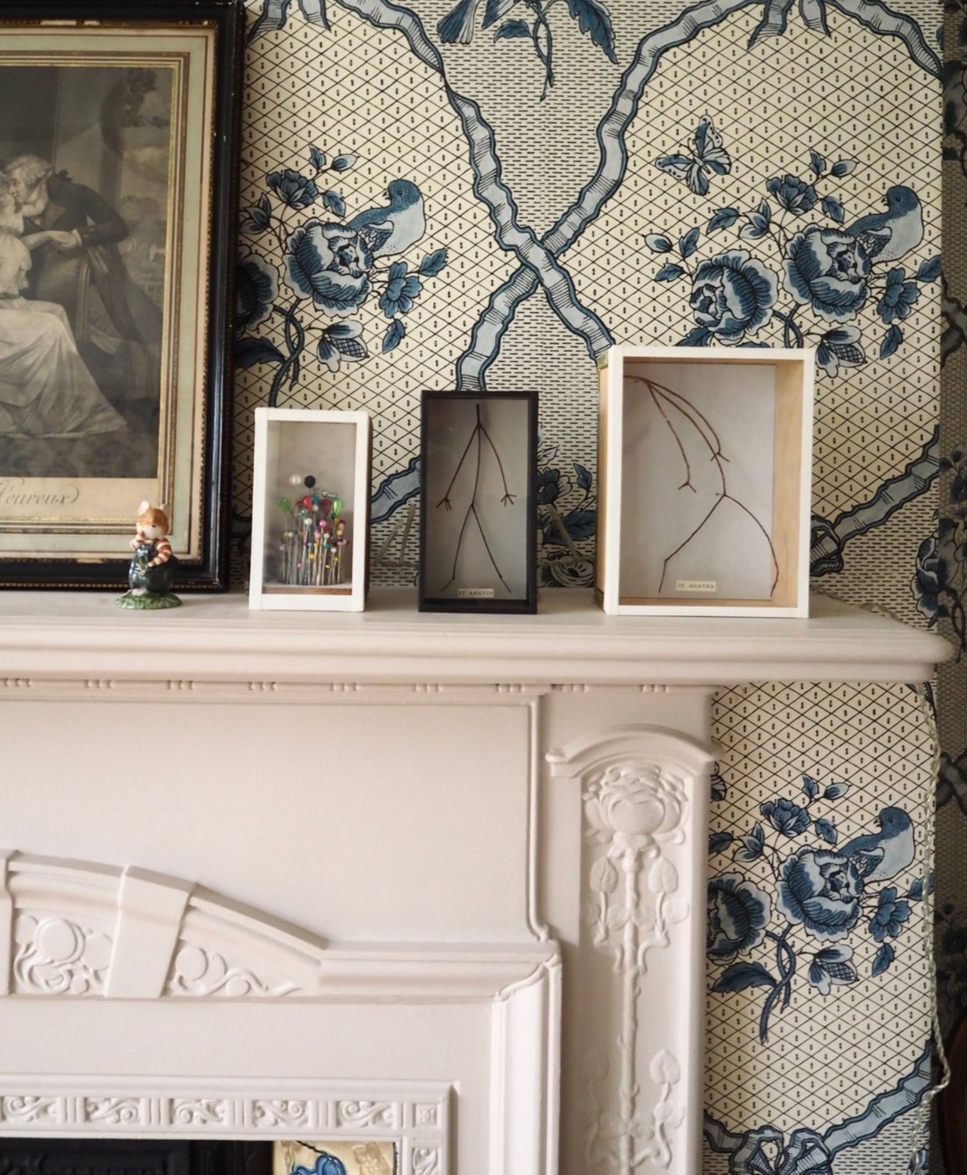 Decorative neutral painted mantelpiece with framed artwork and small sculptures against a patterned wallpaper.