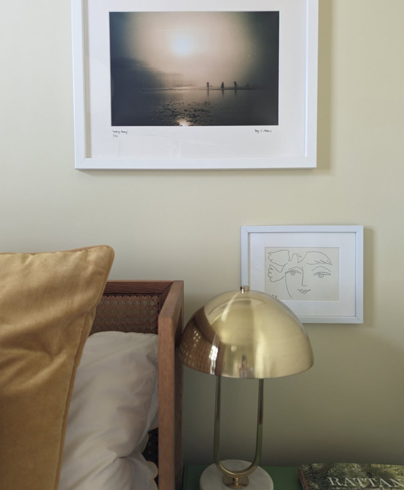 Framed artwork on a buttermilk yellow wall with a gold lamp and cushion in the foreground.