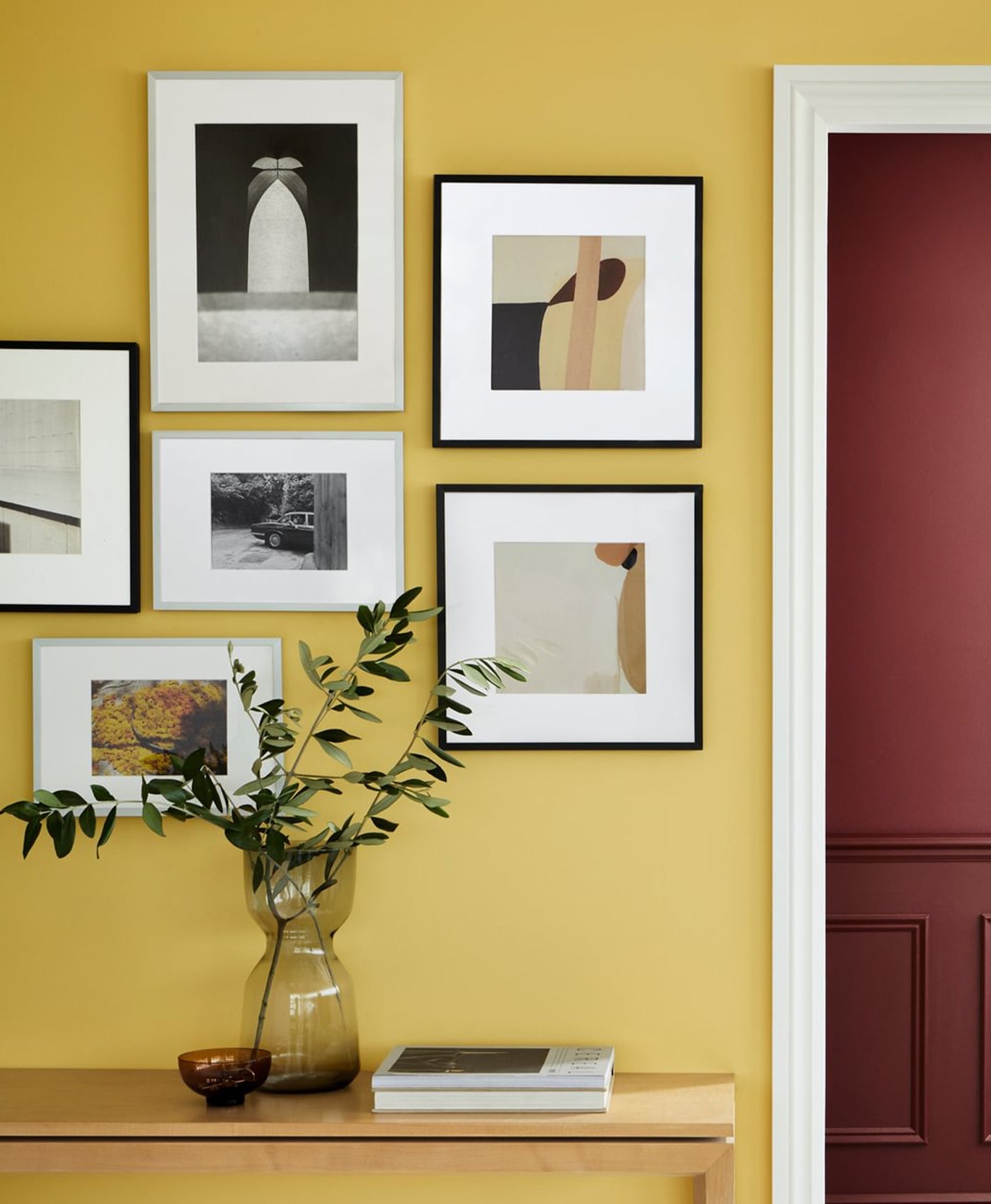 Framed artworks on a sunshine yellow wall with a vase and books on a table.