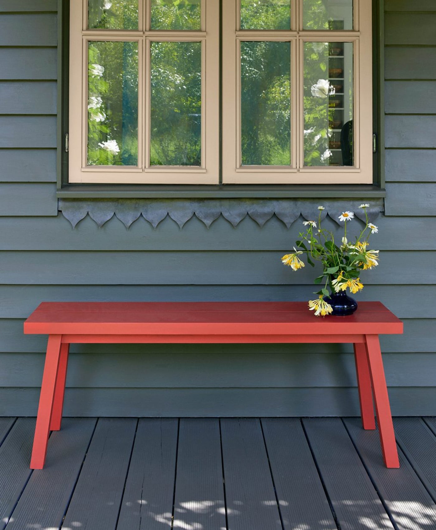 Red bench on a deck with a window and greenery in the background