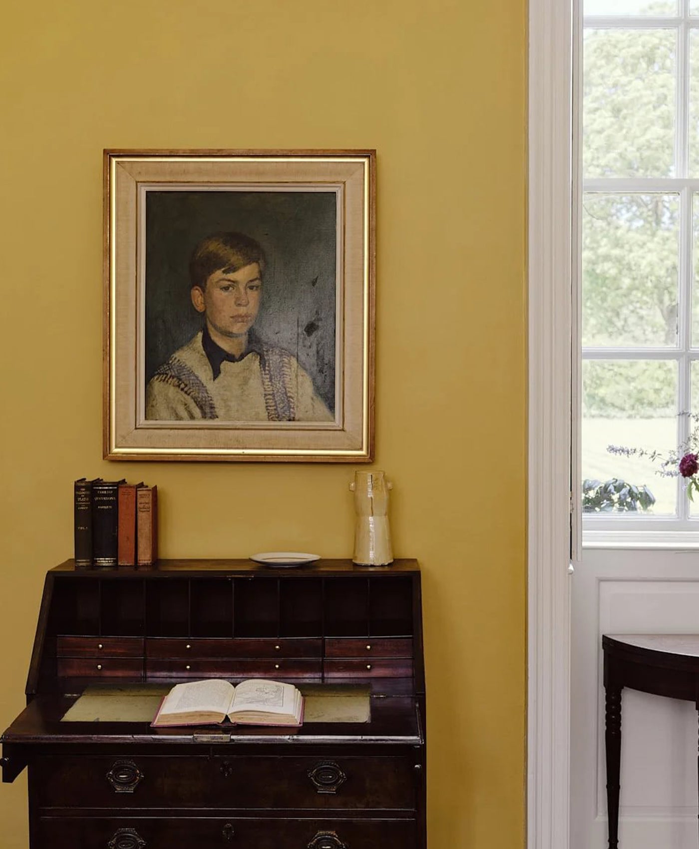 Framed portrait on a yellow wall above a wooden desk with books and an open book.