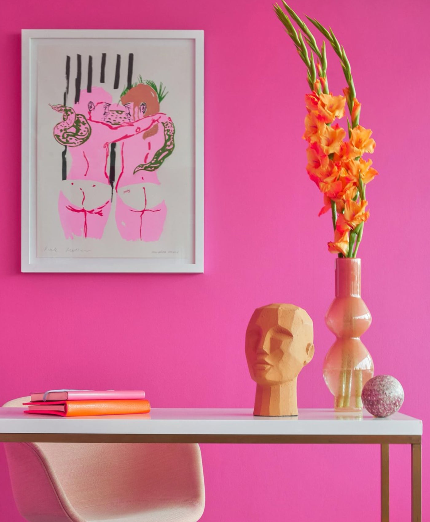 Decorative setup with a framed artwork, vase with flowers, and mannequin head on a table against a bright pink wall.