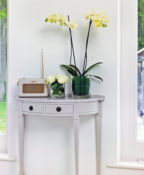 Small lavender table with a radio, glass vase with flowers, and potted plant near a window.