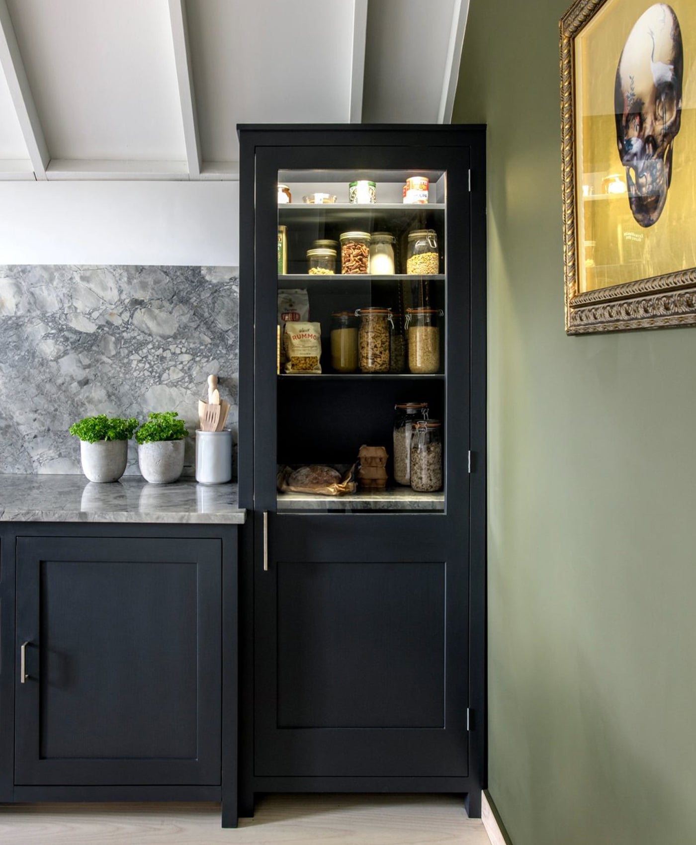Black pantry cabinet with glass door in a kitchen setting, with a green wall and light grey ceiling