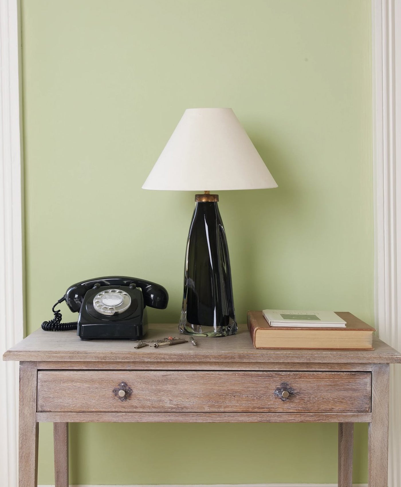 Vintage black telephone, lamp, and books on a wooden side table against a pistachio green wall.