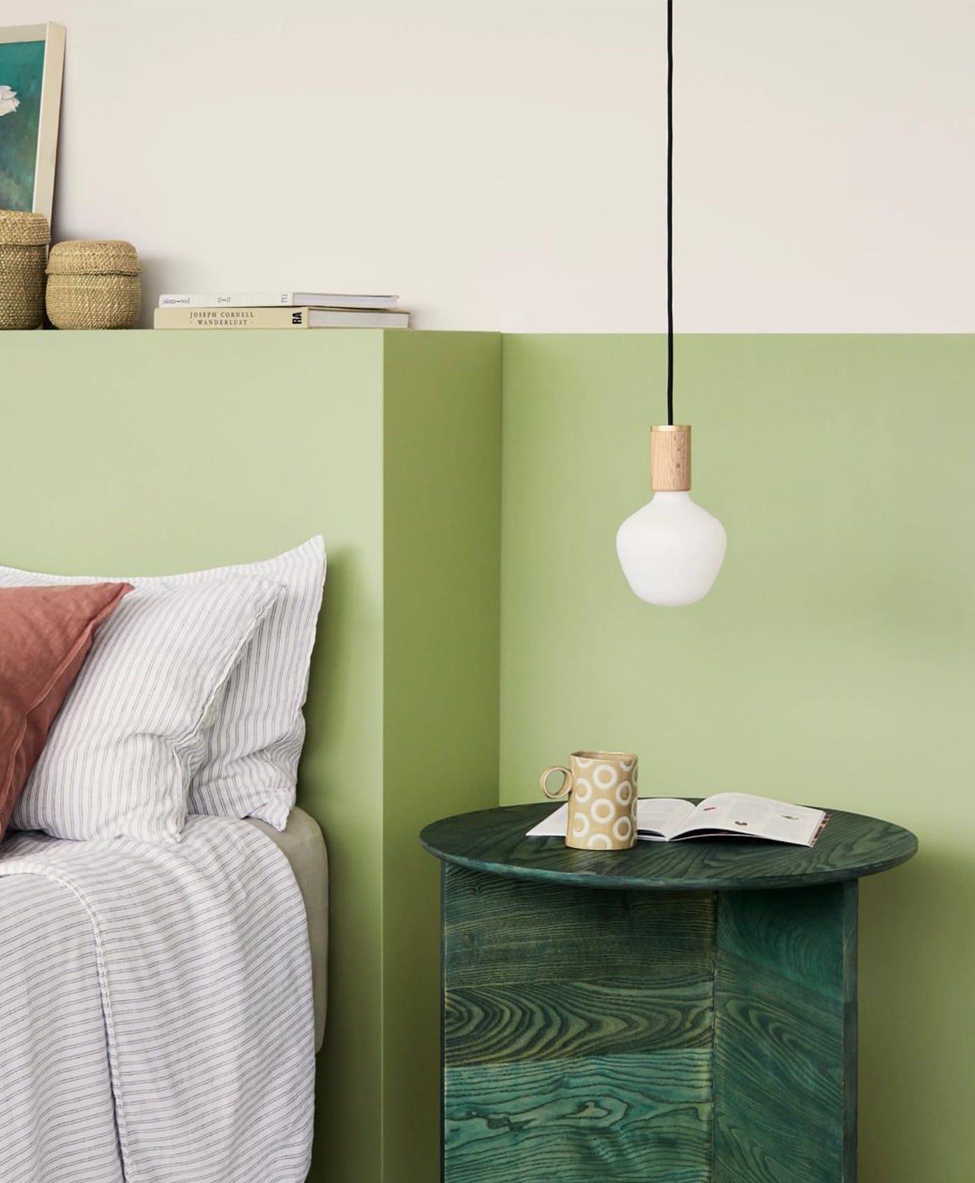 Green side table with a cup and book next to a bed with pillows, against a light green wall.