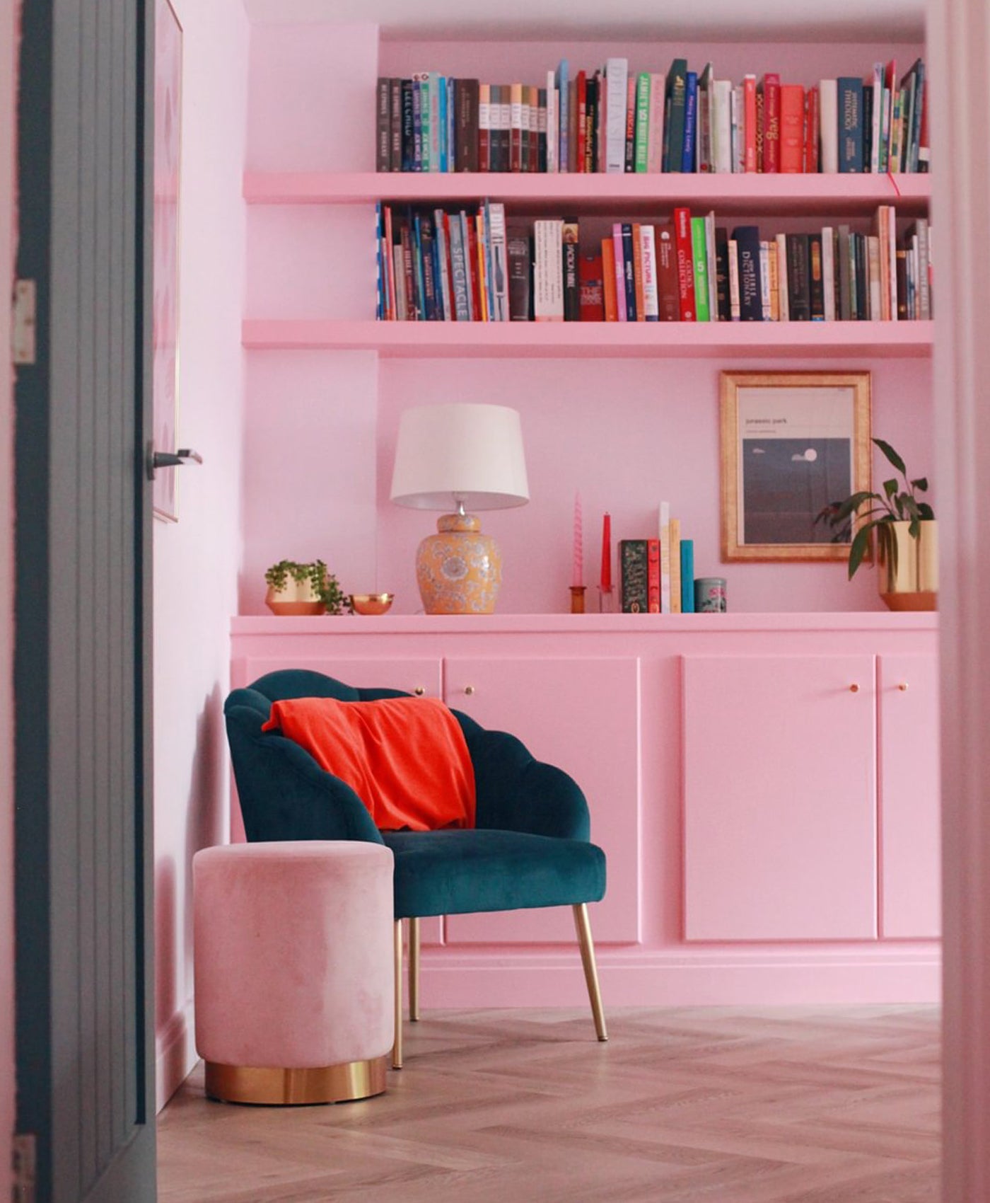 Blush pink room with a teal chair, red cushion, and bookshelf filled with books.