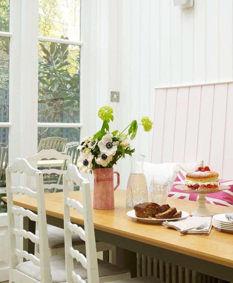 Dining room with a wooden table set for a meal, featuring a cake and flowers and wooden panelled walls in a pale pink colour