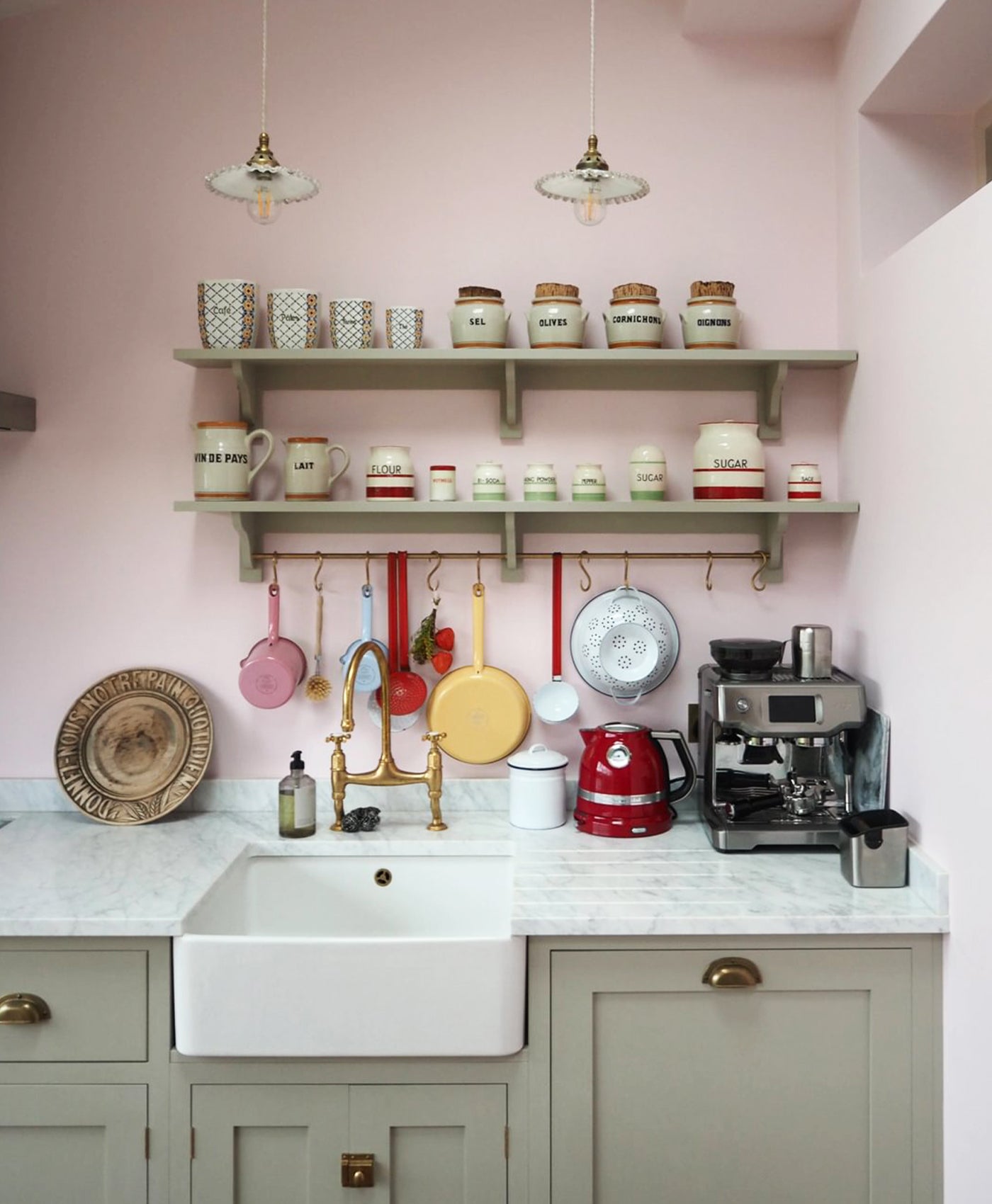 Kitchen with pastel pink walls, shelves with jars and pots, and a white sink.
