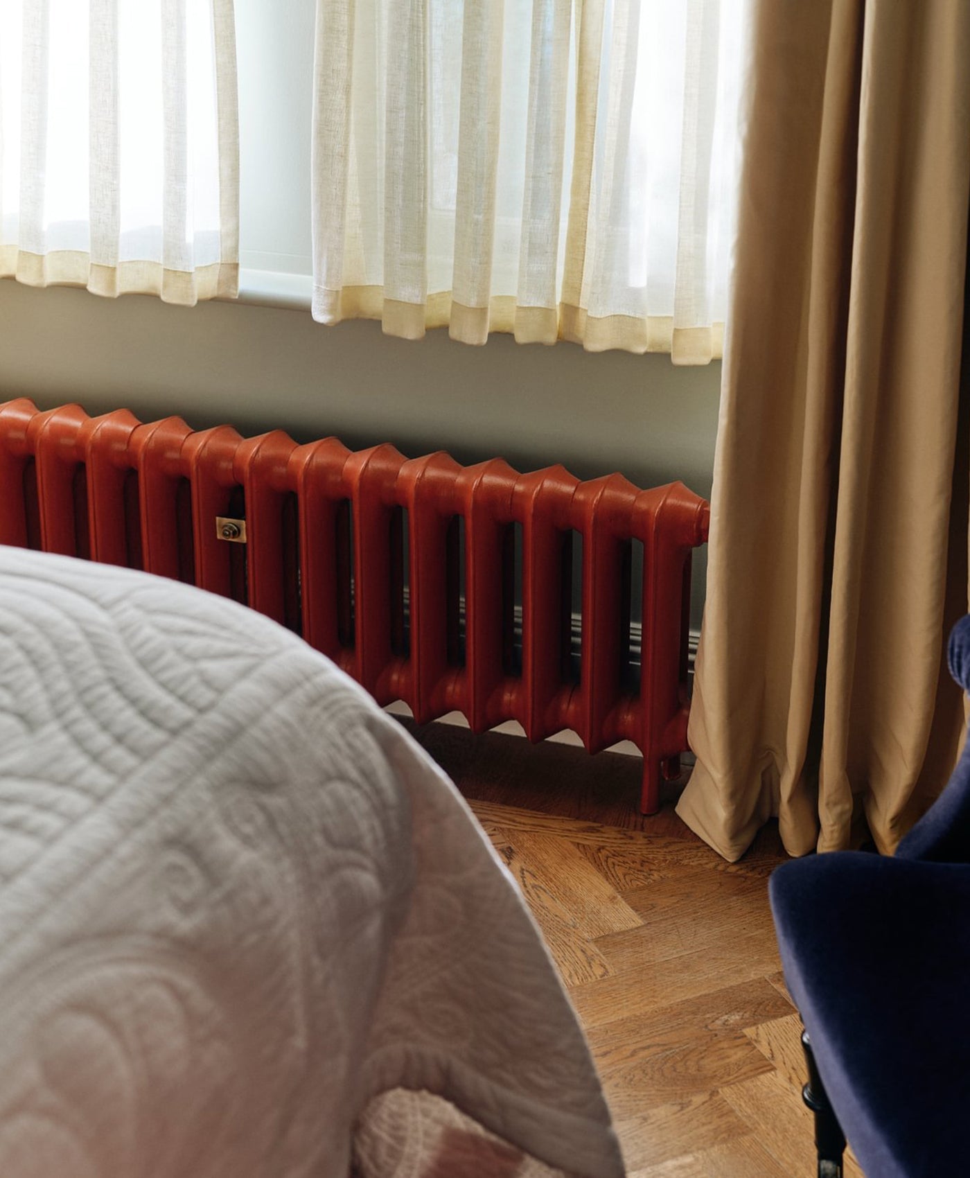 Red radiator against a wall with curtains and a bed in the foreground