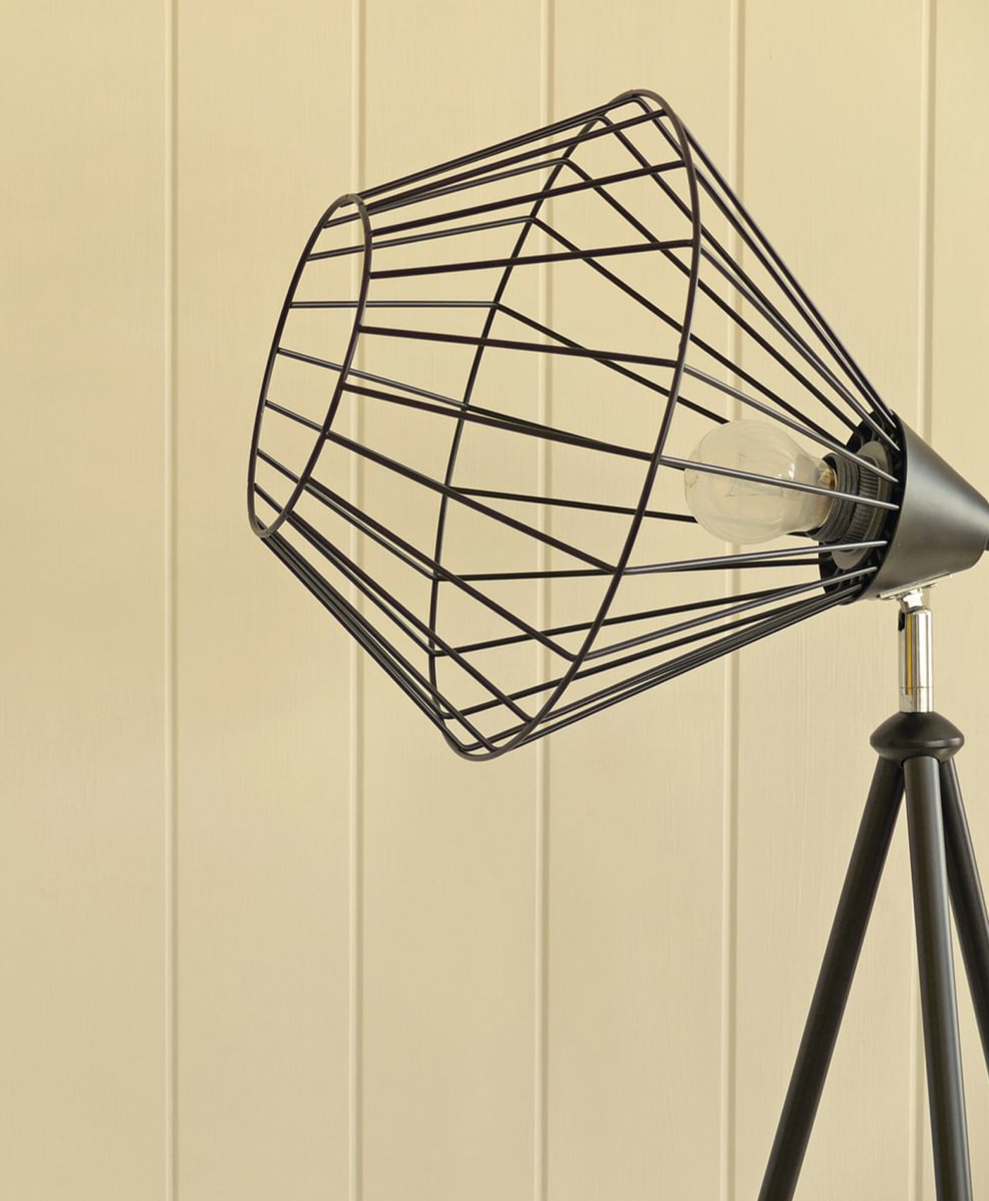Black wire lampshade on a tripod stand against a butter yellow wall.