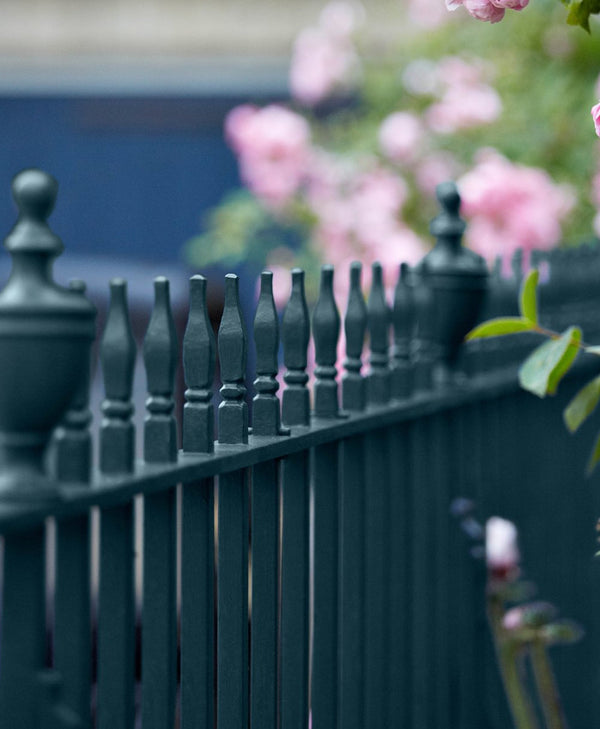 Blue-black metal fence with decorative top rail and blurred pink flowers in the background