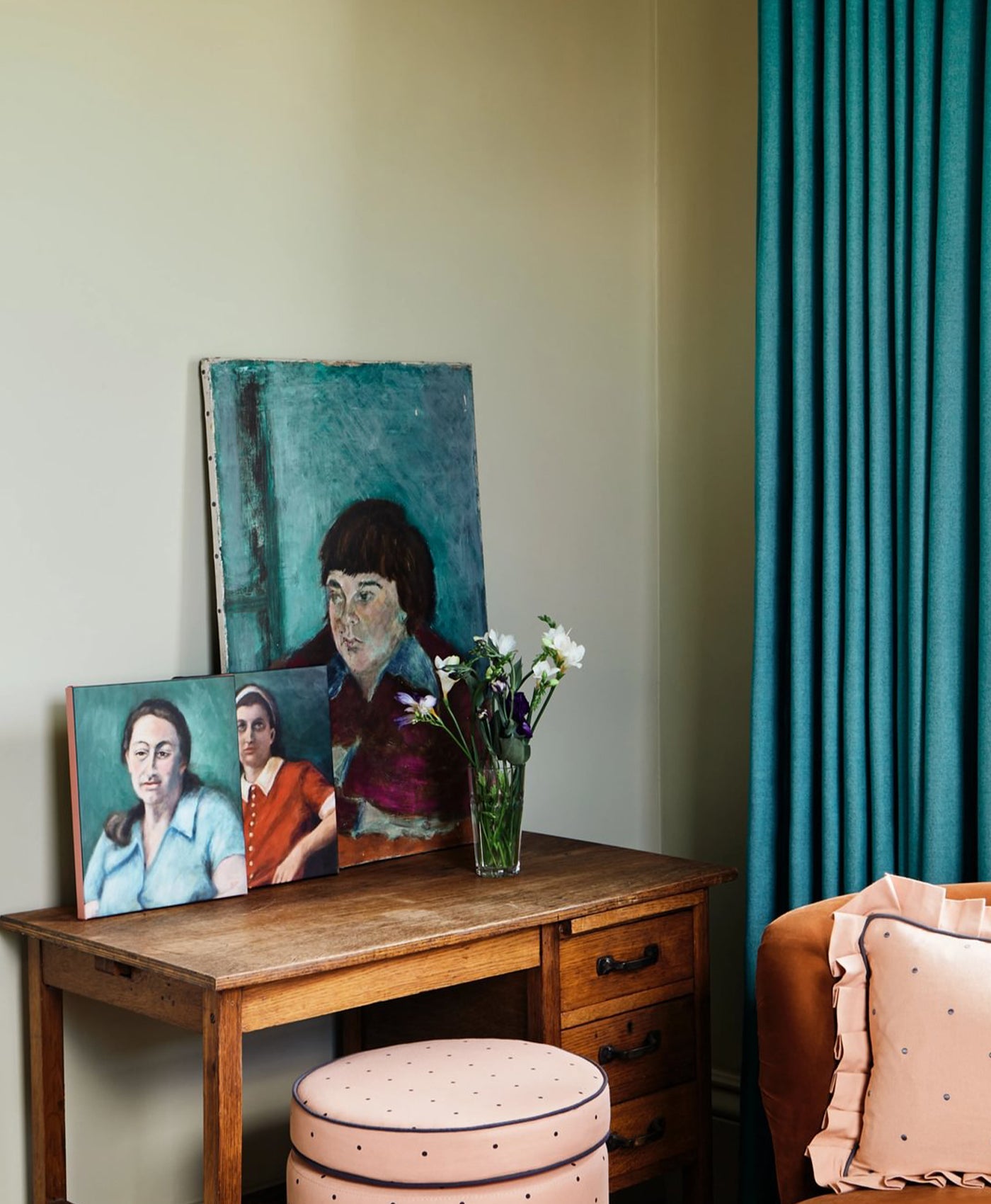 Wooden desk with portraits and a vase of flowers against a teal curtain, in room painted in light olive green colour