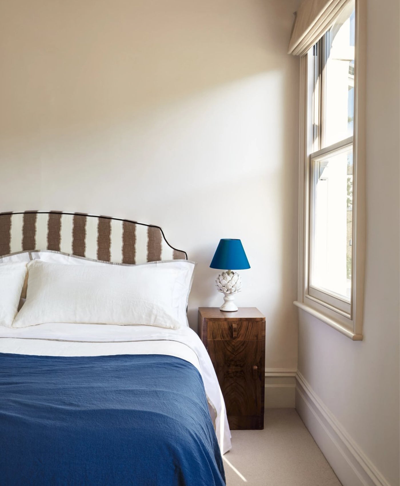 Warm off-white bedroom with striped headboard, blue bedding, and a lamp on a nightstand.