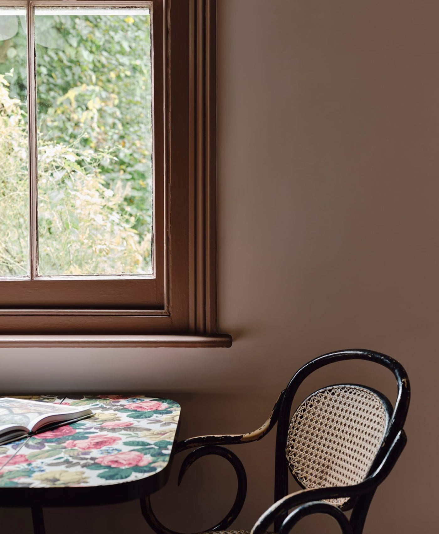 Plaster pink coloured room. Small table with floral pattern and chair next to a window with a view of greenery.