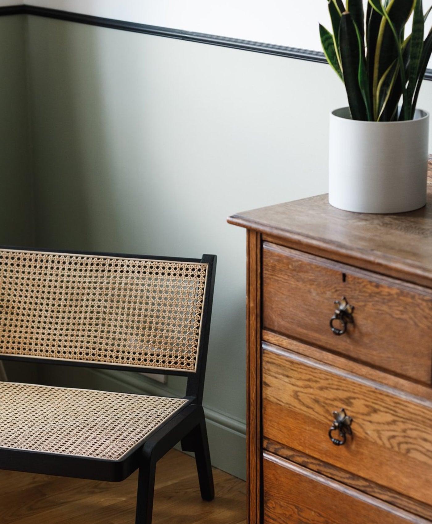 Wooden dresser with a potted plant next to a wicker chair against a light green wall.