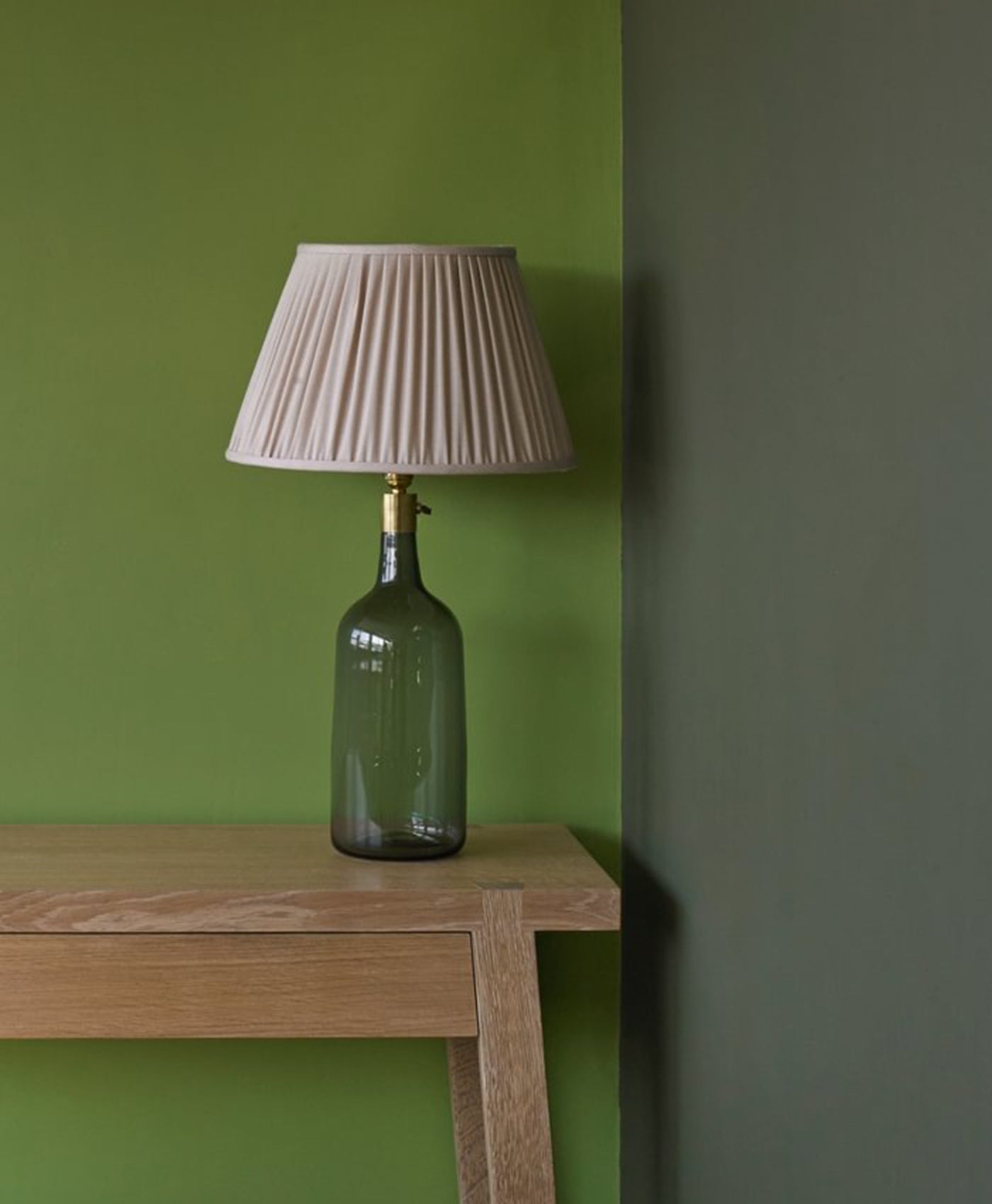 Green bottle lamp with pleated beige shade on a wooden table against an olive green wall