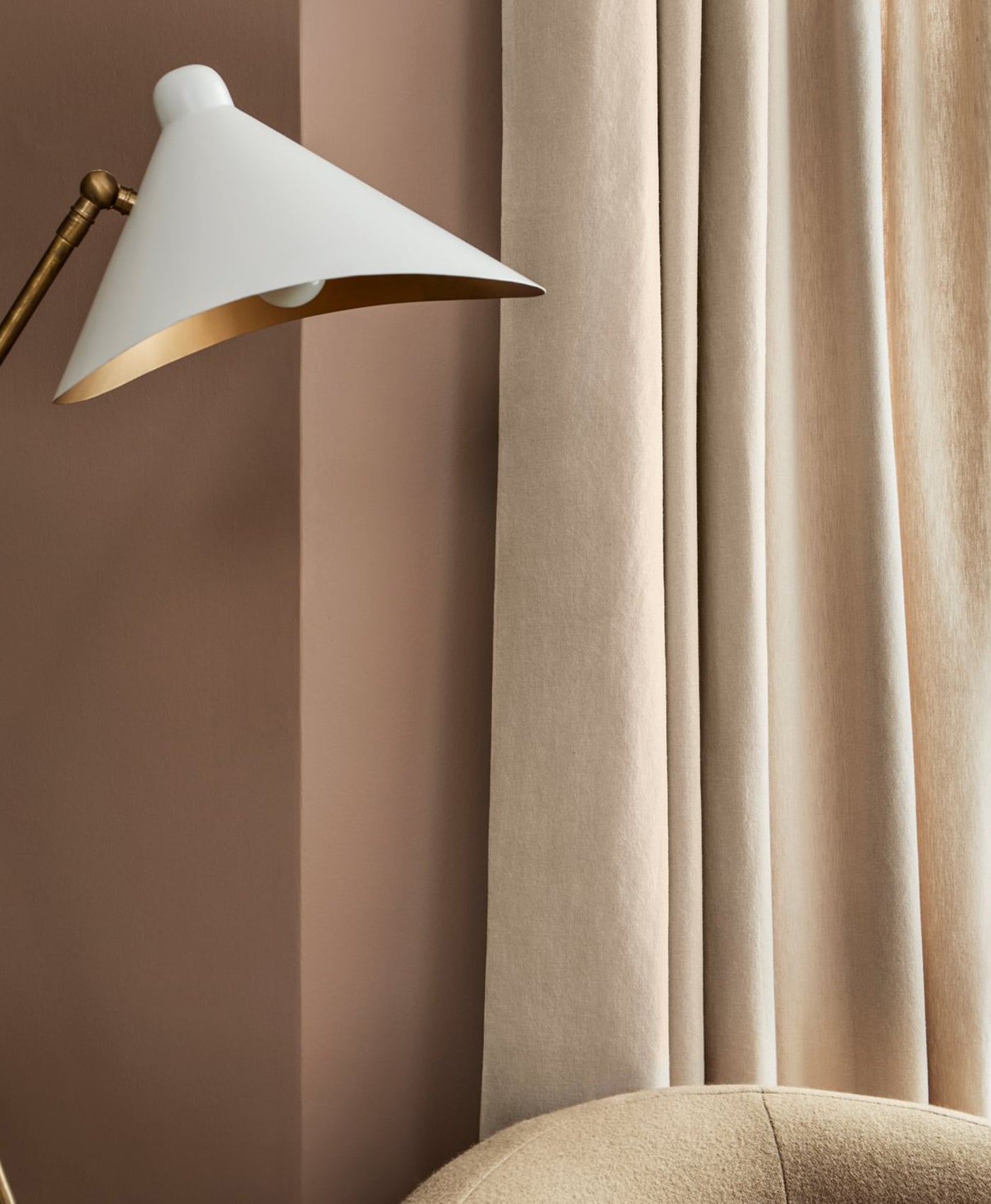 White lamp with gold accents against a beige curtain and plaster pink wall.