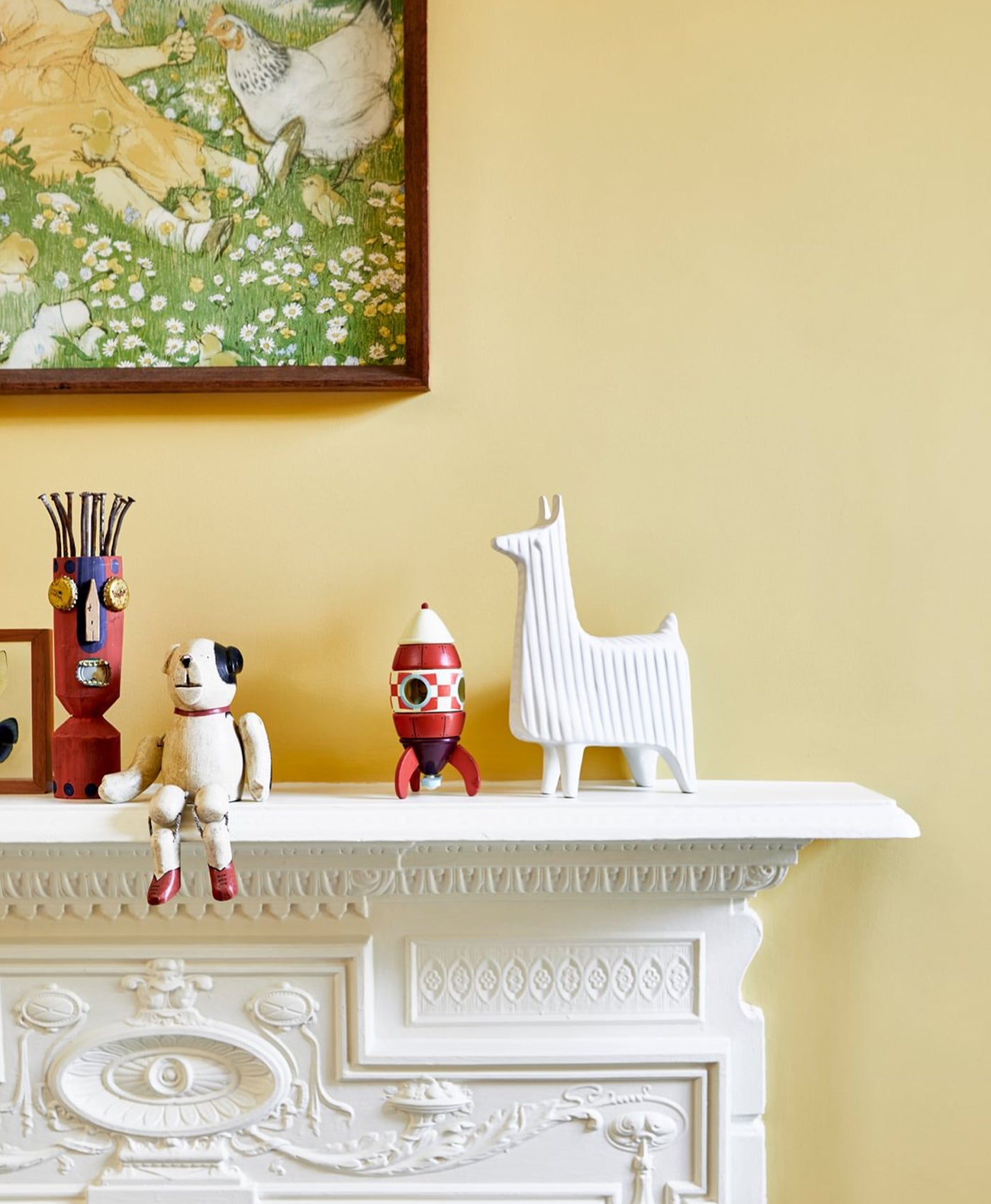 Decorative items on a pale yellow green mantelpiece against a buttercup yellow wall with a framed picture.