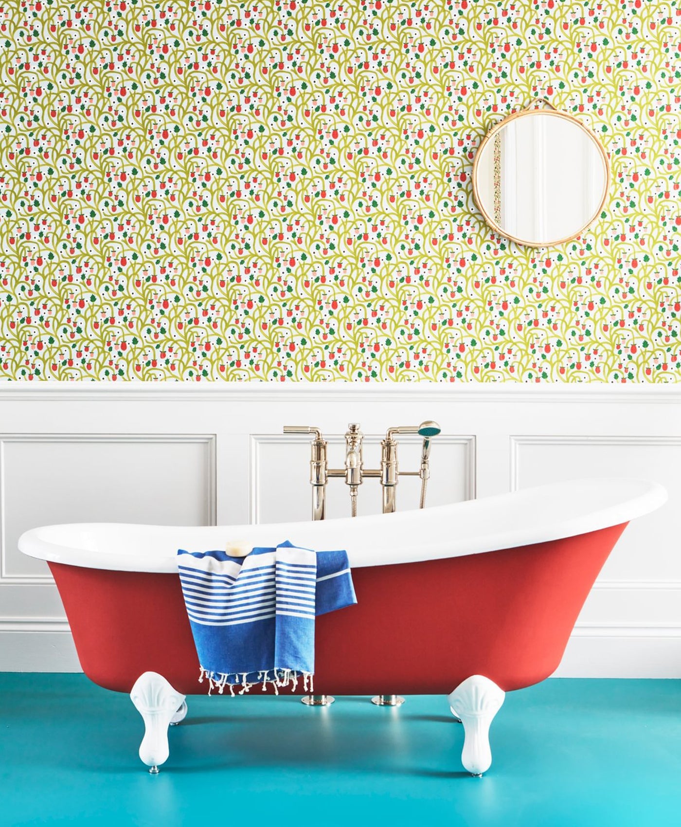 Red bathtub with a blue towel against a colourful floral wallpaper, and white wooden panelling on lower wall.